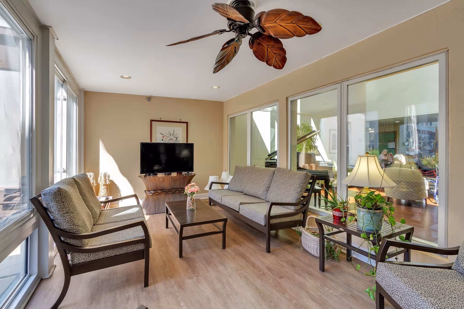 A bright and cozy living room area with large windows on one side allowing natural light to fill the space. The room features a ceiling fan with leaf-shaped blades, a flat-screen TV on a wooden stand, a coffee table with a flower vase, and cushioned wooden chairs and a sofa. There are also potted plants and a table lamp on a side table, with a view into another room through glass windows.