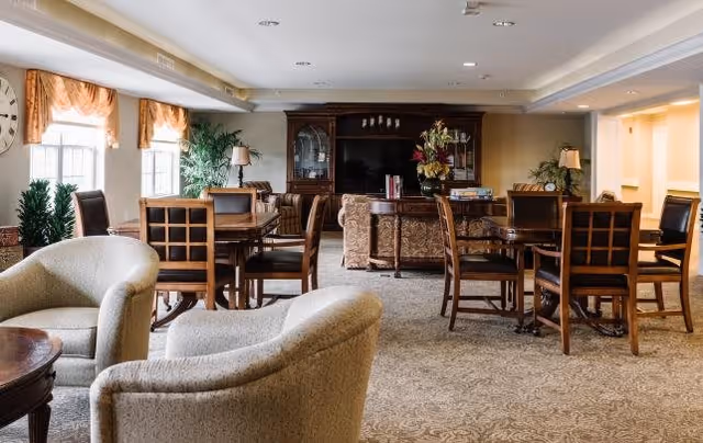 A spacious senior living common area with multiple wooden tables and chairs, beige upholstered armchairs, large windows with curtains, potted plants, and a dark wooden cabinet with decorative items in the background.