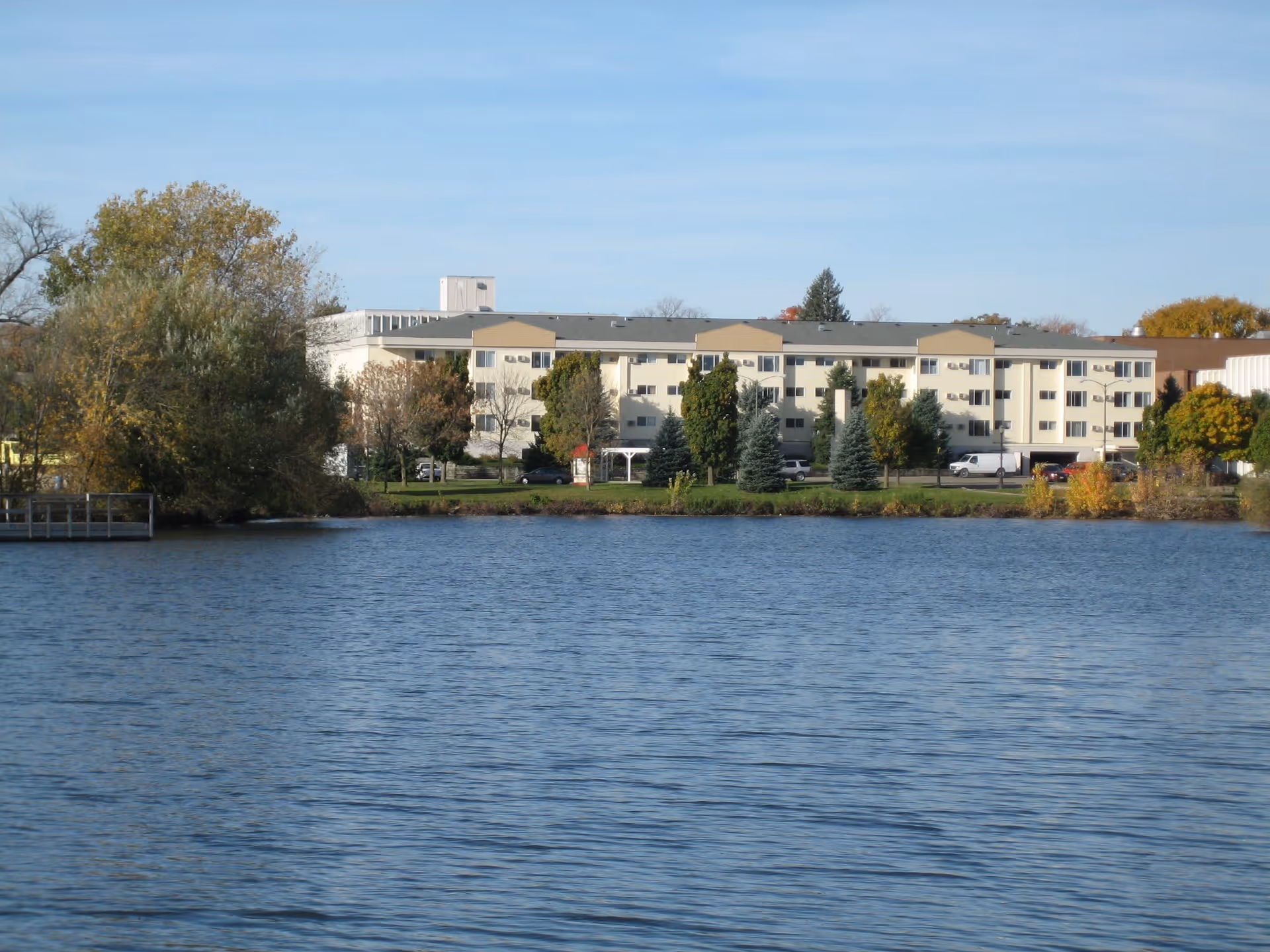 Multi-story senior living building across a lake with trees, lawn, and a small dock in front.