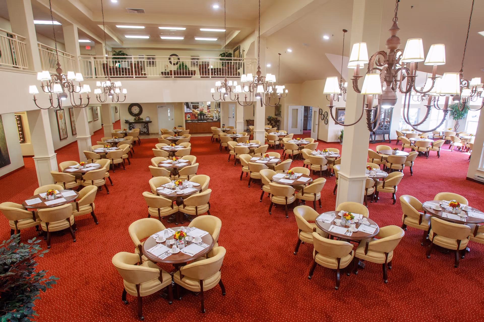 Spacious dining room with multiple round tables each surrounded by four beige cushioned chairs. Tables are set with white napkins, cups, and small floral centerpieces. The room features red carpet flooring, cream-colored walls, large chandeliers hanging from the ceiling, and a second-floor balcony overlooking the area.