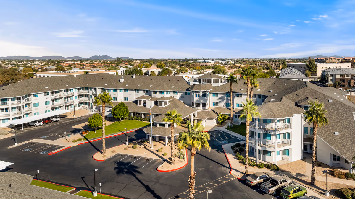 Aerial view of Vista del Rio - A Provincial Senior Living Community showing a large, multi-story building with a gray roof and white exterior walls. The building is surrounded by palm trees, landscaped areas, and a parking lot with several cars. The background includes a suburban area with houses and distant mountains under a blue sky with some clouds.