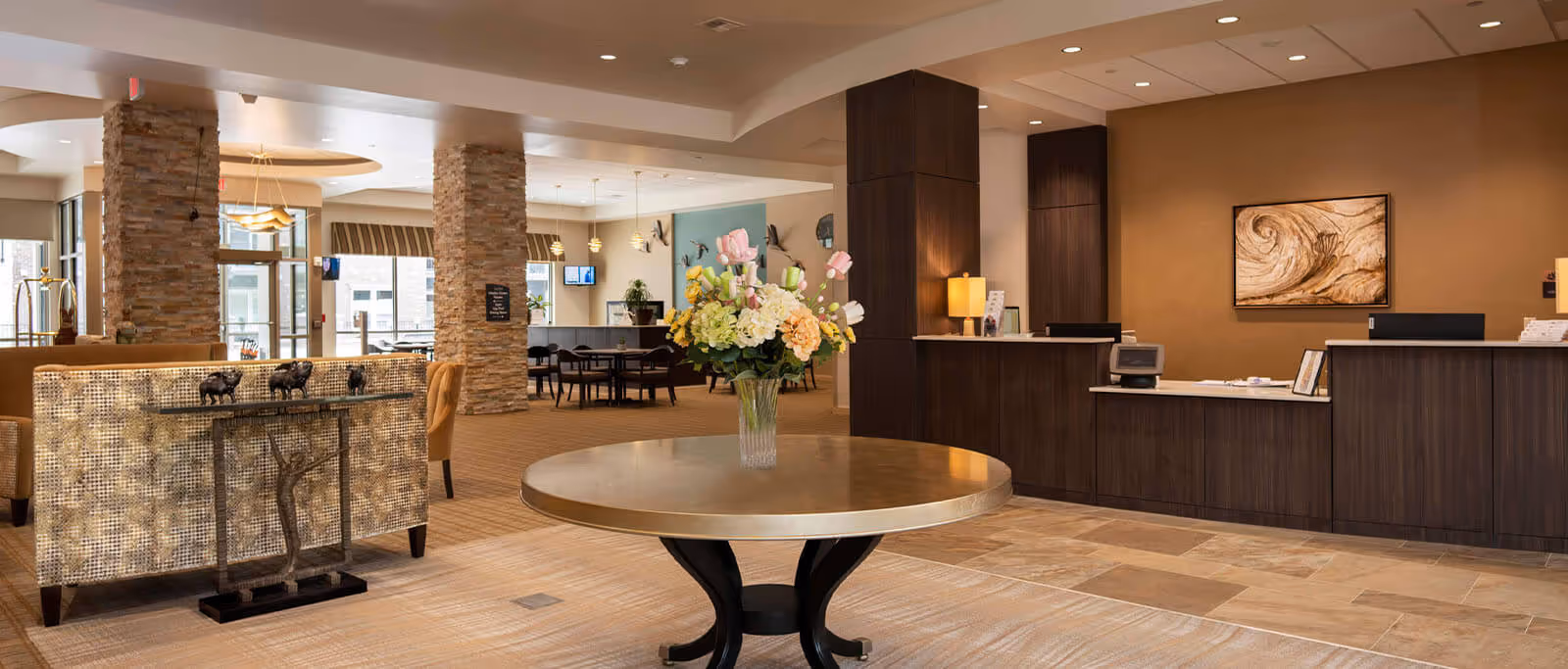 A spacious and well-lit senior living facility lobby with a round table in the center holding a vase of colorful flowers. To the right is a reception desk with dark wood paneling and a lamp, and behind it is a wall with a framed abstract artwork. The left side features comfortable seating and stone pillars, with large windows allowing natural light to fill the space. In the background, there are dining tables and chairs near more windows.