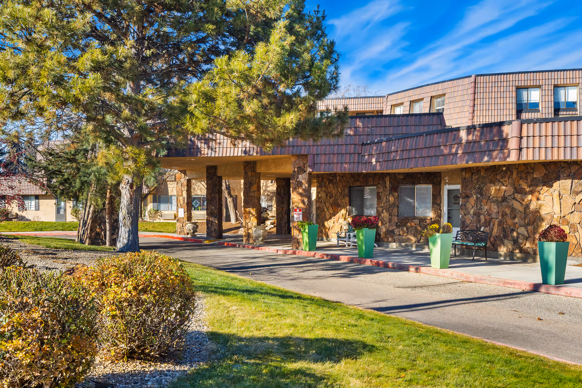 Front entrance of a stone-clad senior living building with a covered driveway, green planters, benches and trees on a sunny day.