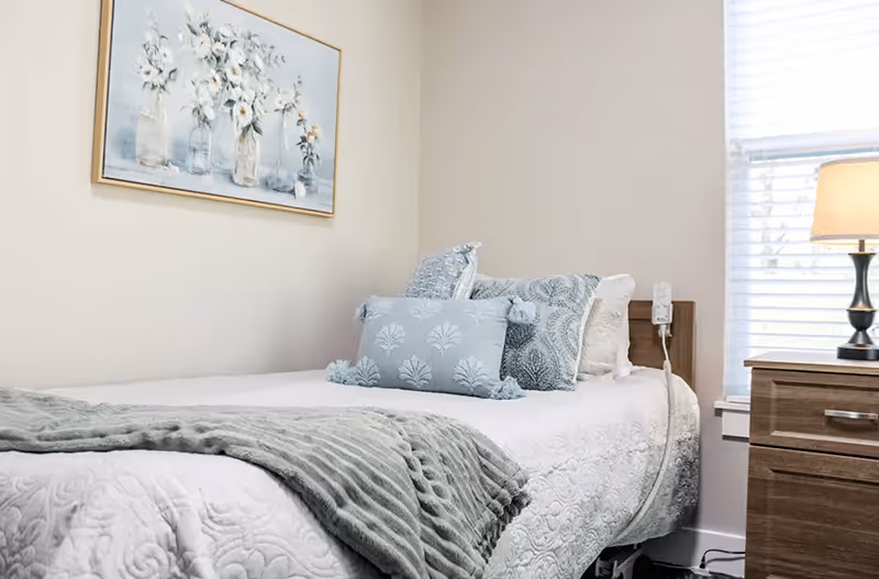 Single bed in a softly lit bedroom with blue-gray pillows and throw, a nightstand with a lamp, and a floral wall painting.