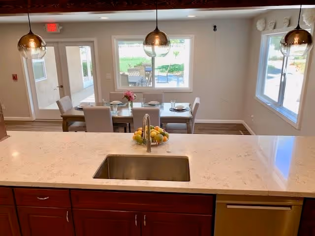 View from a kitchen island with a sink and a bowl of fruit looking out towards a dining area with a table set for four and large windows showing an outdoor patio and greenery.