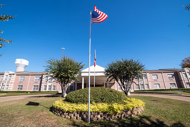 Front exterior view of The Eastman Senior Living facility with a circular landscaped area featuring a flagpole flying the American flag and another flag, two trees, and a water tower in the background under a clear blue sky.