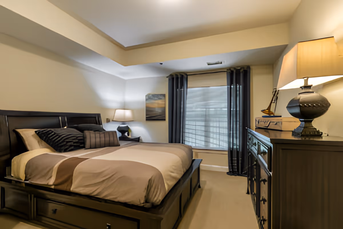A neatly made bed with brown and beige bedding in a bedroom. The room features a dark wooden bed frame, a nightstand with a lamp, a dresser with a lamp and decorative items, a window with blinds and dark curtains, and a piece of wall art.