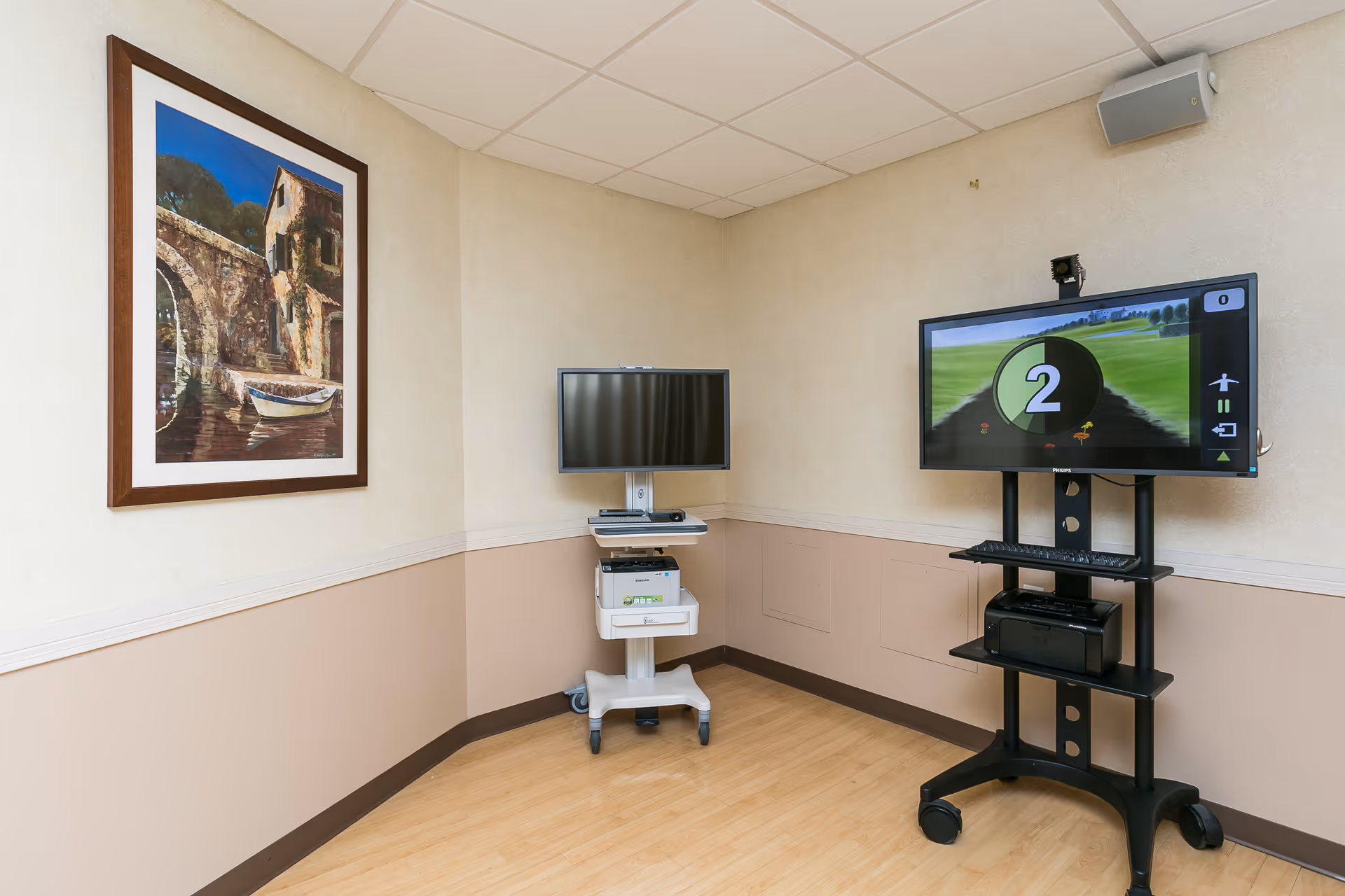 A corner of a room with two flat-screen monitors on mobile stands, one displaying a game with a large number 2 on the screen. The walls are beige with a chair rail and a framed painting of a stone bridge and boat on the left wall. The floor is light wood.