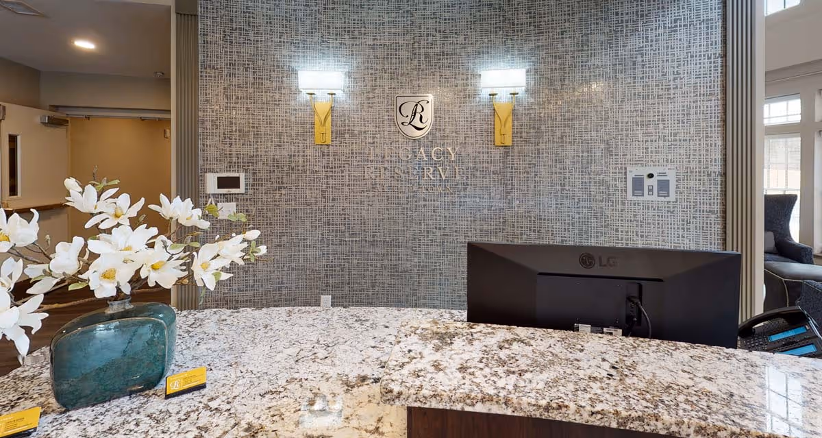 Reception desk area at Legacy Reserve at Old Town with a granite countertop, a green vase with white flowers, a computer monitor, and a textured wall behind featuring the Legacy Reserve logo and two wall sconces.