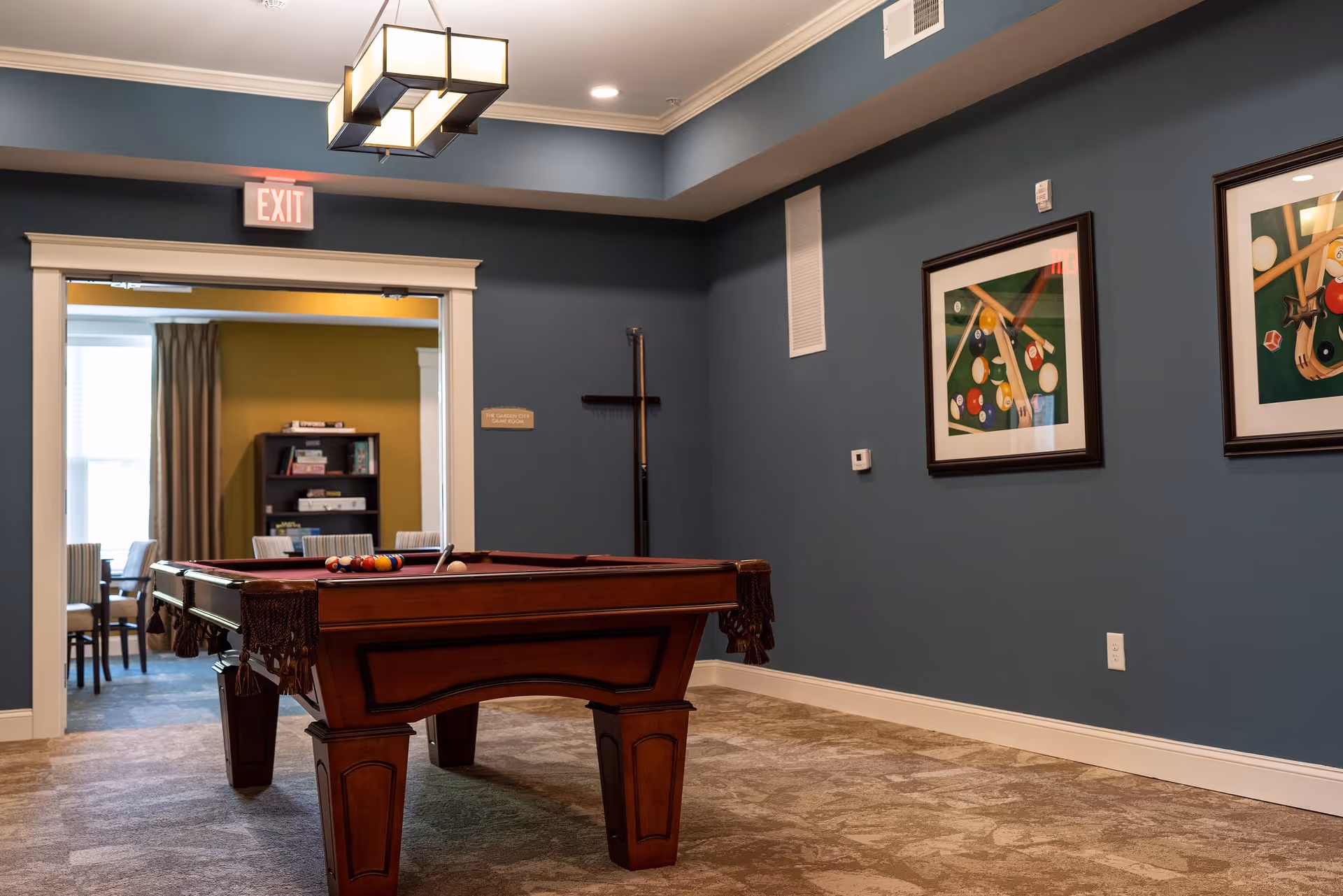 Interior room with a pool table in the center, blue walls adorned with framed pictures of billiard balls and cues, a ceiling light fixture, and an exit sign above a doorway leading to another room with a bookshelf and chairs.