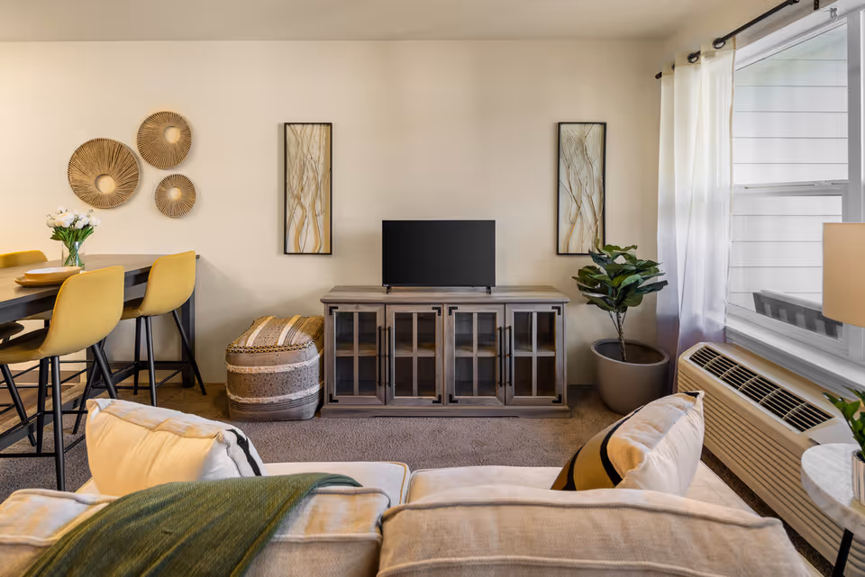 A cozy living room area with a beige sofa in the foreground, a wooden TV stand with a flat-screen TV, two framed artworks on the wall, a potted plant, and a window with white curtains. To the left, there is a dining table with yellow chairs and decorative wall hangings above it.