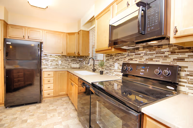A bright kitchen with light wood cabinets, a black refrigerator, black microwave, black electric stove, and a black dishwasher. The backsplash features a mosaic tile design in neutral tones, and there is a window with white blinds above the sink. The floor has a patterned tile design in beige tones.