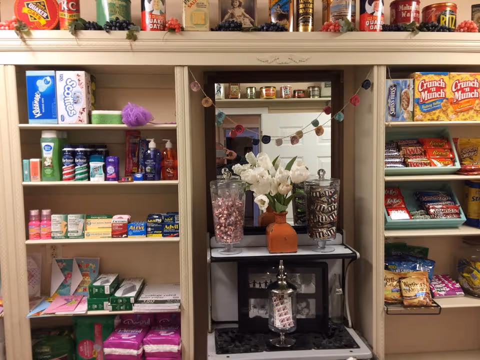 Shelves stocked with various personal care items, snacks, and medications. The left shelves contain hygiene products, over-the-counter medicines, and feminine hygiene products. The right shelves display snacks like Crunch n Munch, candy bars, and chips. In the center, there is a mirror with a garland of colorful circular decorations, a vase with white flowers, and two glass jars filled with candy.