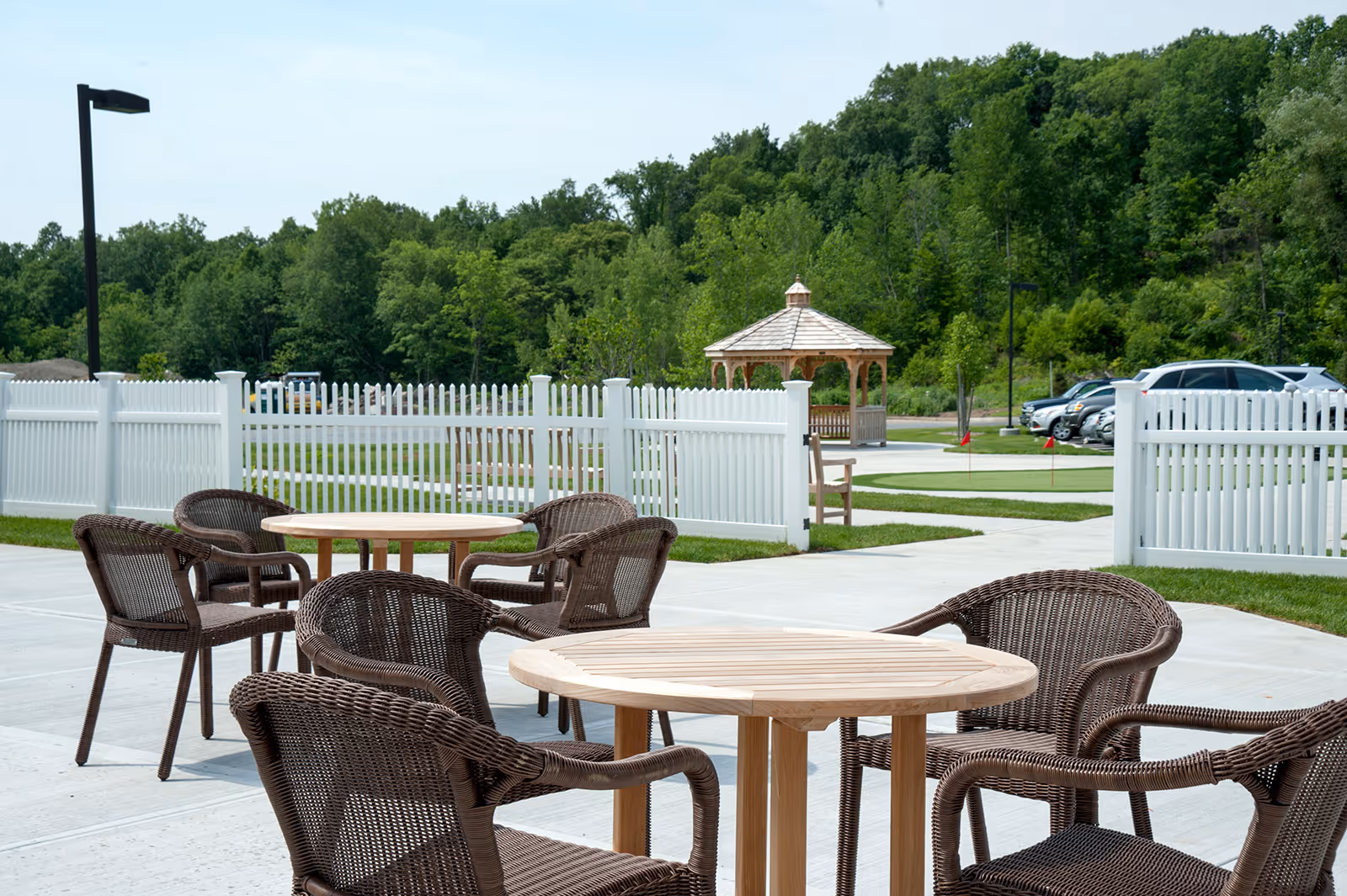 Outdoor patio with round wooden tables and wicker chairs by a white picket fence, gazebo, and parked cars.