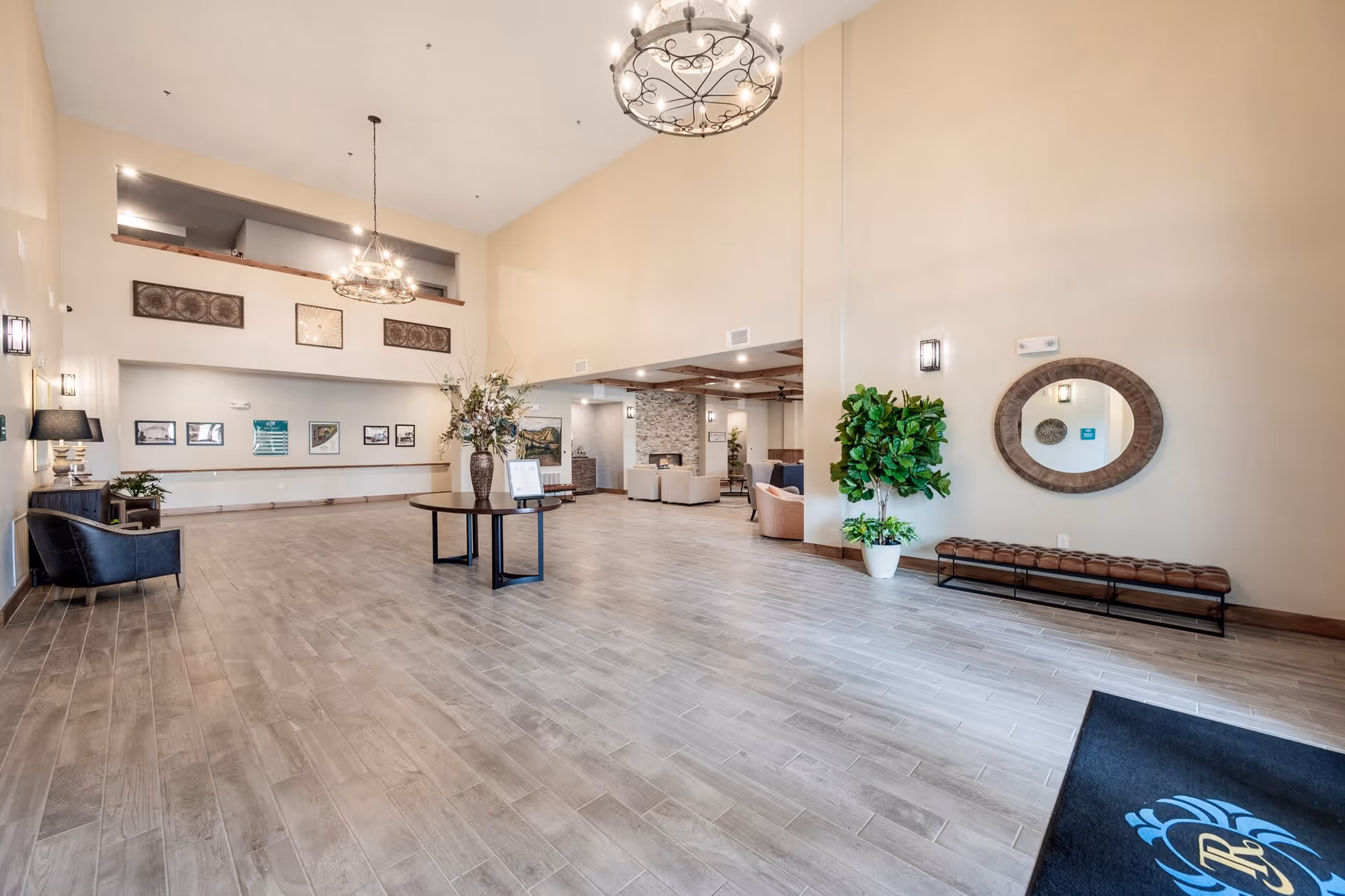 Spacious senior living facility lobby with high ceilings, light-colored walls, wood-look tile flooring, a round table with a floral arrangement in the center, comfortable seating areas in the background, decorative wall art, a large round mirror above a bench, and two elegant chandeliers hanging from the ceiling.