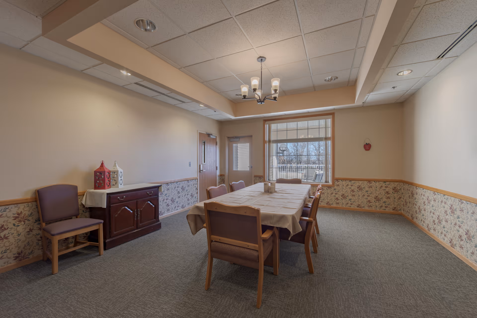A dining room with a rectangular table covered with a beige tablecloth and surrounded by six wooden chairs with padded seats. There is a wooden sideboard against the left wall with two decorative lanterns on top. The walls have floral wallpaper on the lower half and are painted beige on the upper half. A window and a door are visible at the far end of the room, and a modern chandelier hangs from the ceiling.