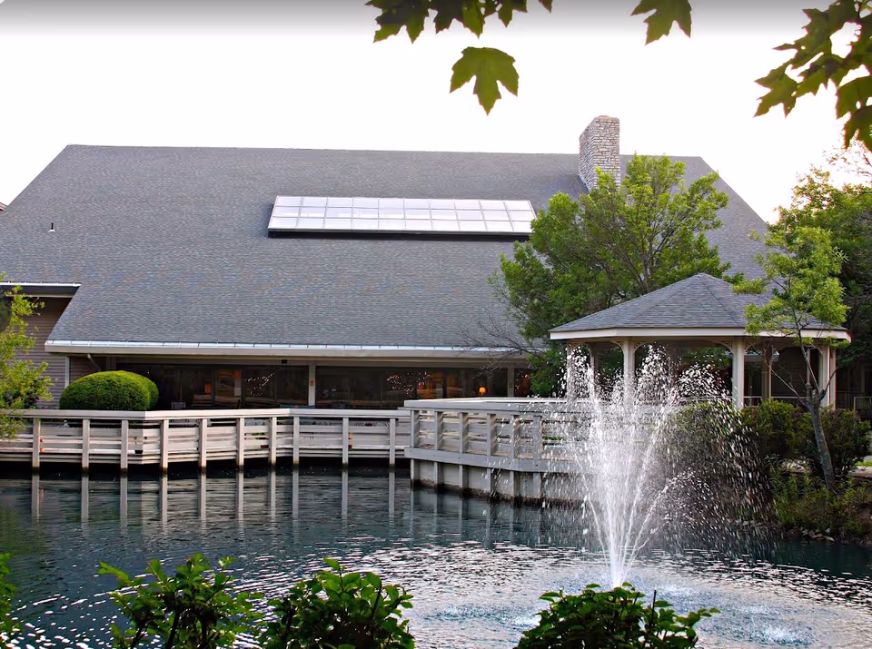 A pond with a fountain, wooden boardwalk and gazebo in front of a large building surrounded by trees.