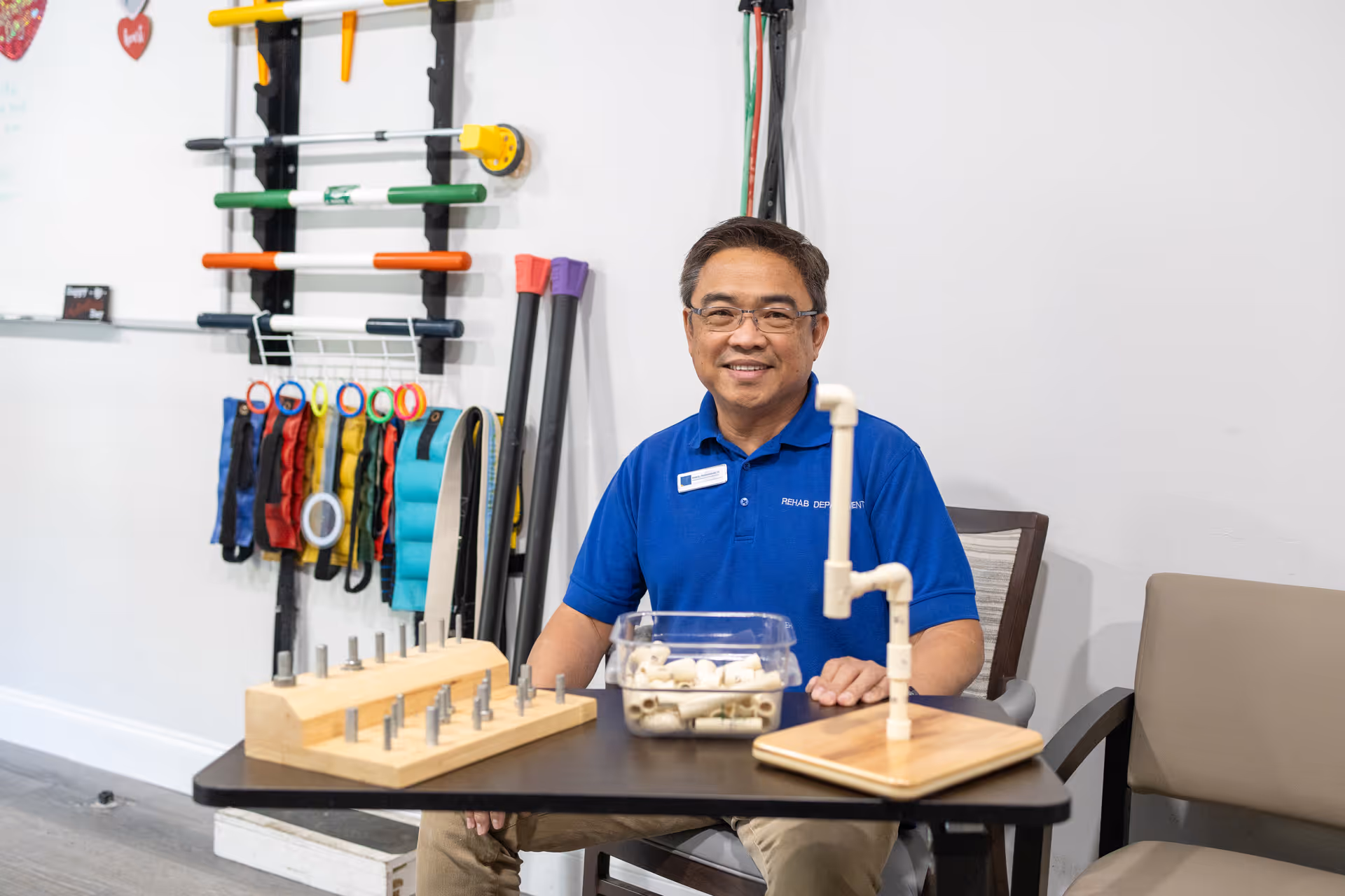 A man wearing glasses and a blue Rehab Department polo shirt sits at a table with various rehabilitation tools and equipment. Behind him on the wall are colorful exercise bands and rods organized on racks. The setting appears to be a therapy or rehabilitation room.