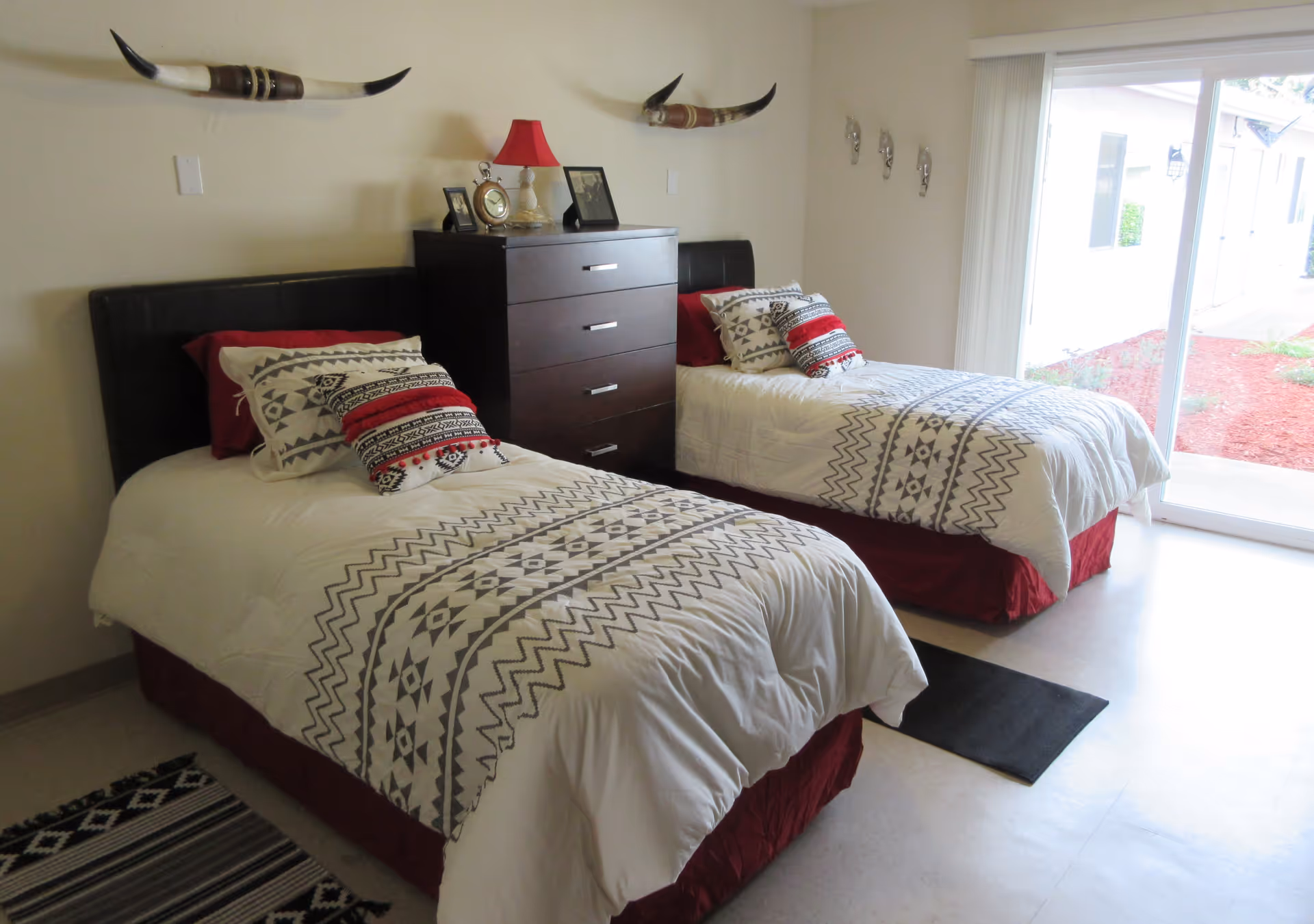 A bedroom with two twin beds featuring white and gray patterned bedding with red accents. Between the beds is a dark wooden dresser with a red lamp, a clock, and framed photos on top. Above each bed is a decorative longhorn mounted on the wall. A large sliding glass door on the right side lets in natural light and shows a view of the outside.