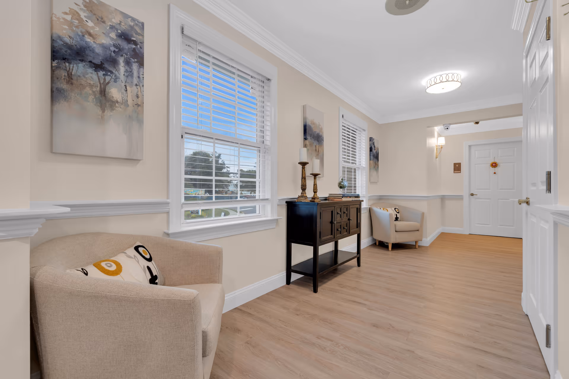 A bright and clean hallway in a senior living facility with light wood flooring, beige walls, and white trim. The hallway features two beige armchairs with decorative pillows, a black console table with candlesticks and books, and three abstract paintings on the walls. Two windows with white blinds allow natural light to enter the space. A white door is visible at the end of the hallway.