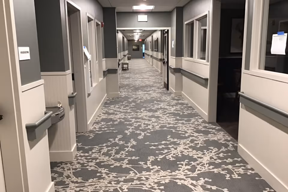A long, well-lit carpeted corridor in a senior living facility with handrails, multiple doorways, and patterned carpeting.