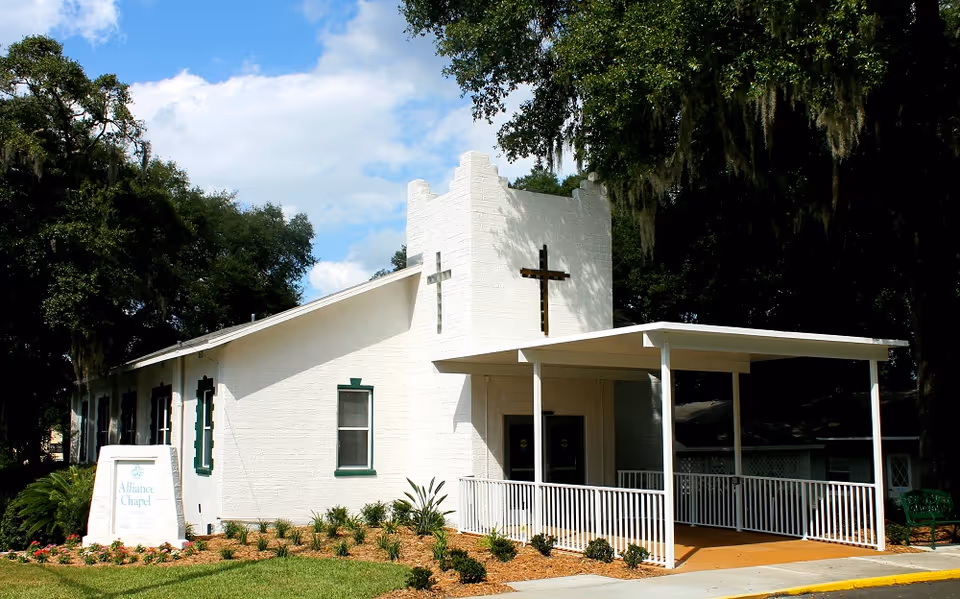 White chapel-style building with two crosses, a covered ramped entrance, and landscaped grounds under large trees.
