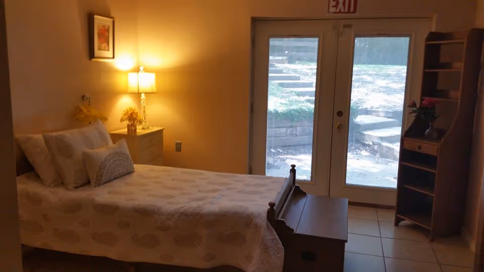 Small, warmly lit bedroom with a single bed, nightstand and lamp beside glass double doors leading outside.