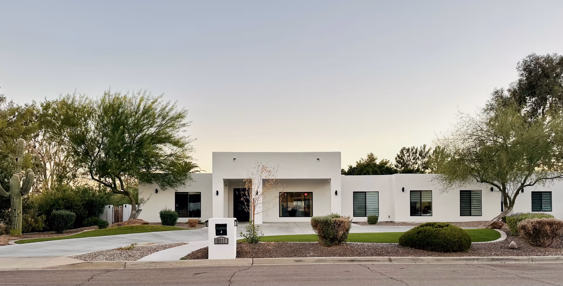 Front exterior view of a single-story white building with a flat roof, surrounded by desert landscaping including trees, bushes, and a large cactus. A concrete driveway and walkway lead to the entrance, and a white mailbox with the number 8111 is visible in front.