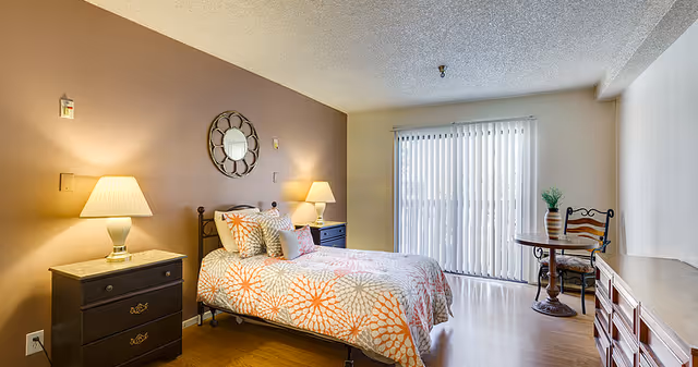 A well-lit bedroom with a bed featuring an orange and white patterned bedspread, two bedside tables with lamps, a round decorative mirror on the wall, a small round table with a chair near a sliding glass door with vertical blinds, and a wooden dresser.