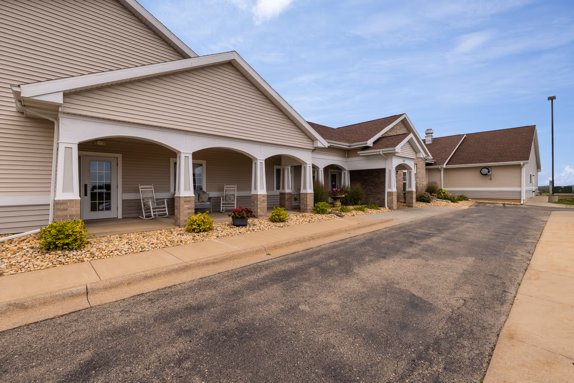 Exterior view of Clover Ridge Place showing a beige building with a covered porch area featuring white columns and rocking chairs. There are small bushes and flower pots along the edge of the porch, and a paved driveway runs alongside the building under a partly cloudy sky.