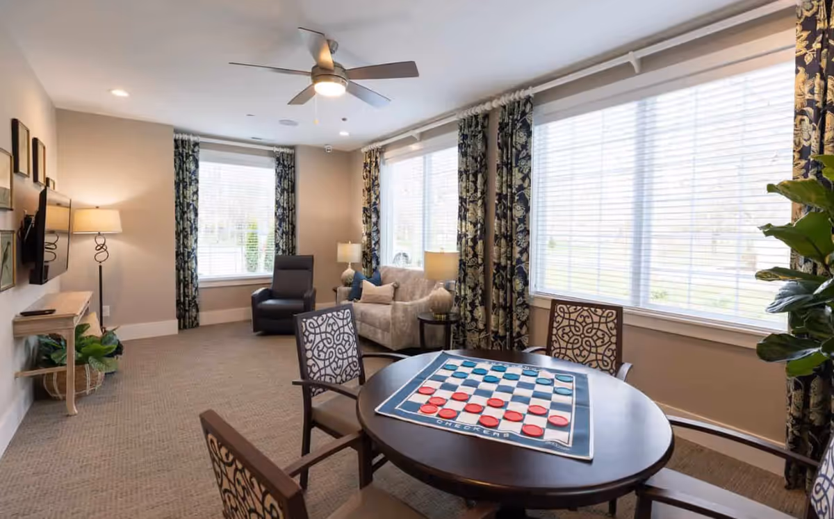 A bright and cozy living room with large windows covered by patterned curtains. The room features a round table with a checkers game set up, surrounded by four chairs with decorative backs. There is a beige sofa with pillows, a dark armchair, two table lamps, a ceiling fan with light, a wall-mounted TV, and a small wooden console table with a plant underneath.