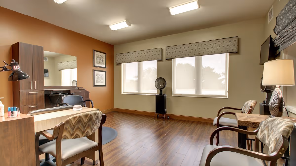 A well-lit room with wooden flooring featuring a vanity table with a chair, a large mirror, and a salon-style chair. Two windows with patterned valances allow natural light to enter. There are two additional chairs with patterned upholstery and a small table with a lamp. A hair dryer on a stand is positioned near the windows.
