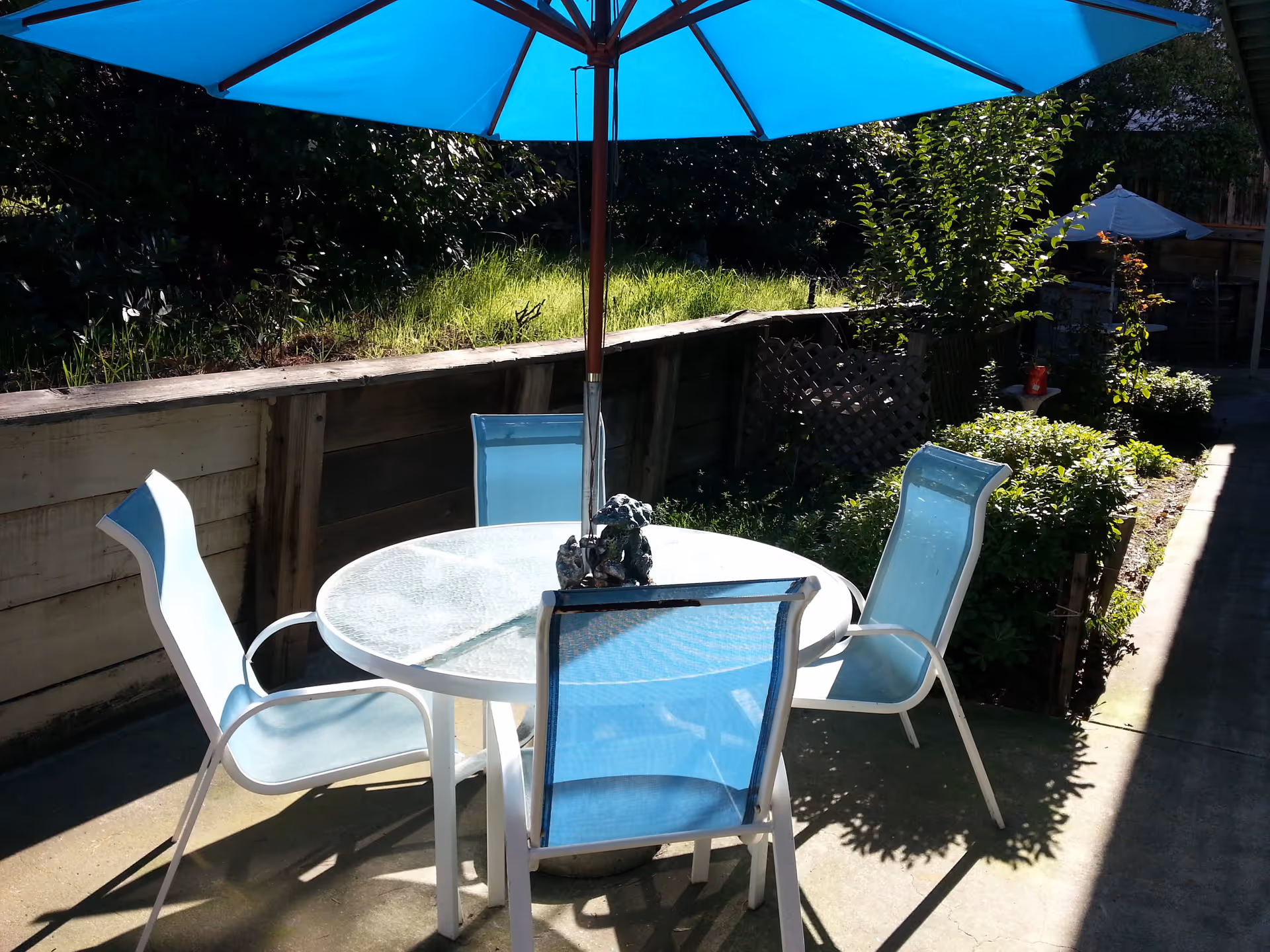 Outdoor patio with a round glass table, four light-blue chairs and a large blue umbrella next to a garden bed.
