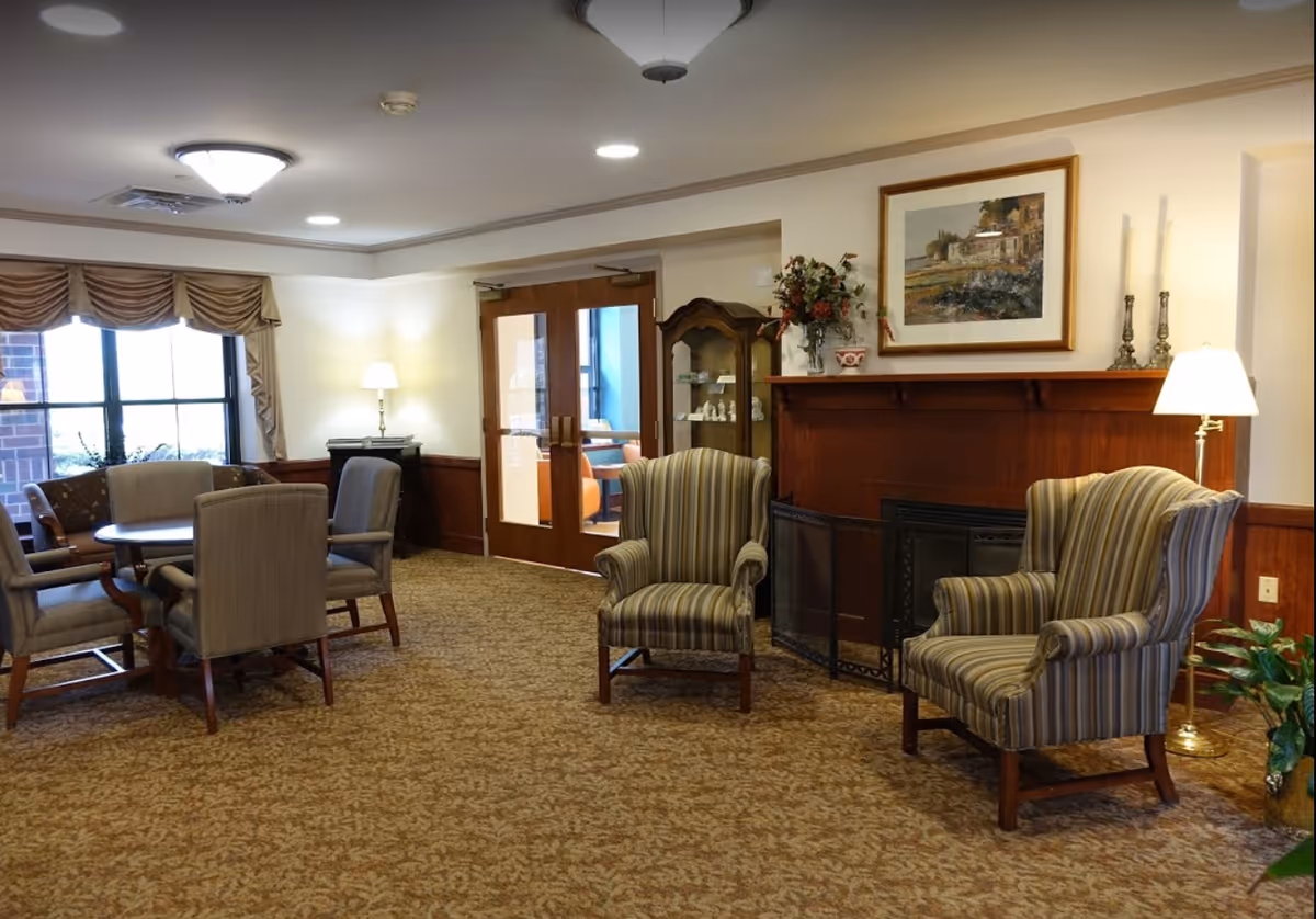 Common sitting room in a senior living facility with upholstered armchairs, a round table and chairs, a fireplace and lamps.