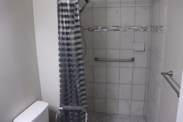 A bathroom featuring a tiled walk-in shower with a grab bar and a soap holder. The shower has a striped shower curtain. A toilet is partially visible on the left side, and a towel bar is mounted on the right wall.