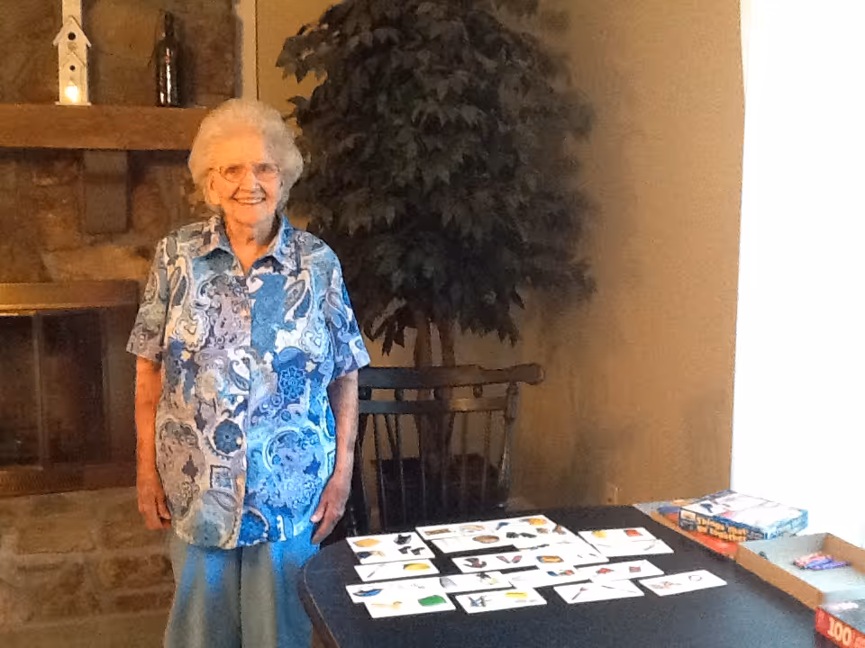 An elderly woman with white hair and glasses stands smiling next to a table with various picture cards spread out on it. Behind her is a stone fireplace with decorative items on the mantel and a large potted plant. The setting appears to be a cozy indoor room.
