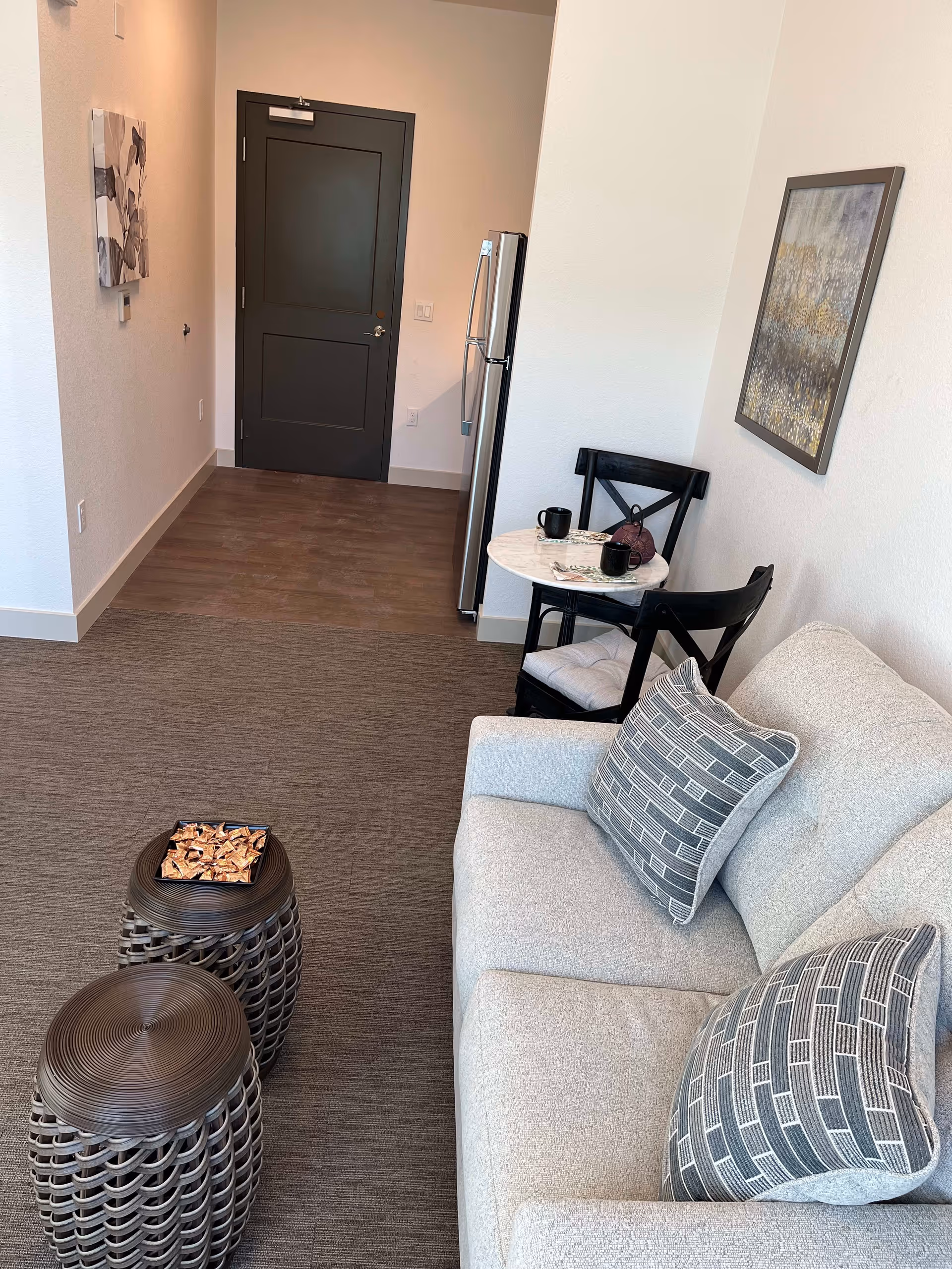A small living area with a light gray couch featuring two patterned pillows, two dark woven side tables with a decorative bowl on one, a small round table with two black chairs, two black mugs, and a teapot. In the background, there is a stainless steel refrigerator and a closed dark door. The walls are white with a framed abstract painting and a floral wall art.