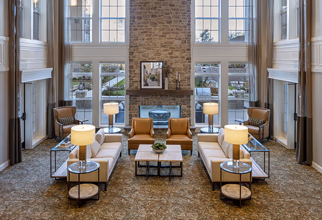 A spacious and elegant senior living facility common area with high ceilings and large windows. The room features a stone fireplace in the center with a framed picture above it. There are two white sofas facing each other with a wooden coffee table in between, flanked by two brown leather armchairs. Four table lamps with white shades are placed on round side tables next to the sofas. The carpet has a patterned design, and tall curtains frame the windows.