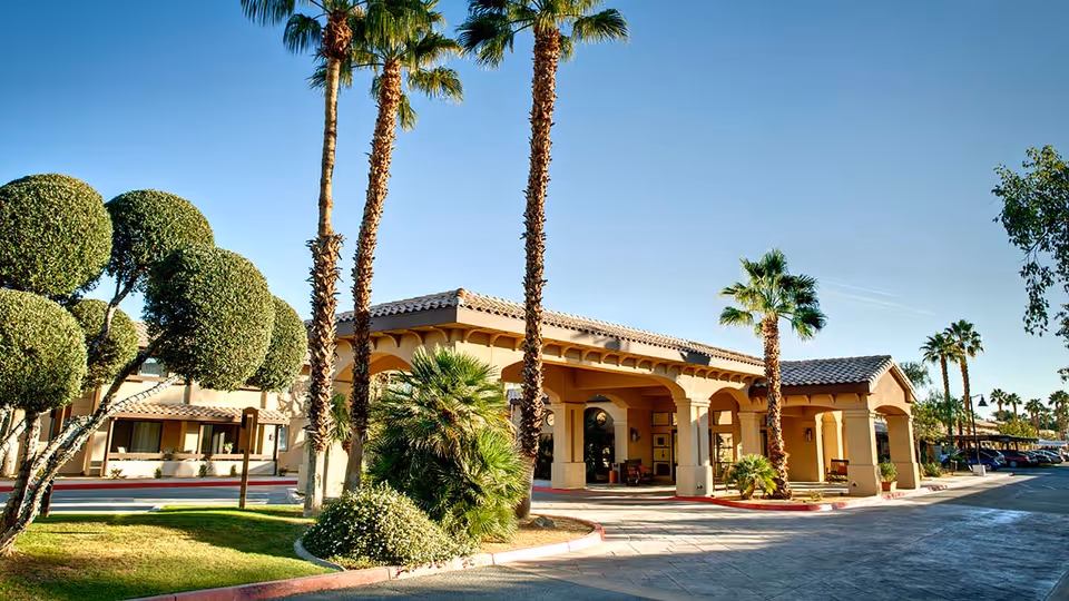 Exterior view of Atria Hacienda senior living facility featuring a Mediterranean-style building with arched entrances, palm trees, manicured bushes, and a clear blue sky.