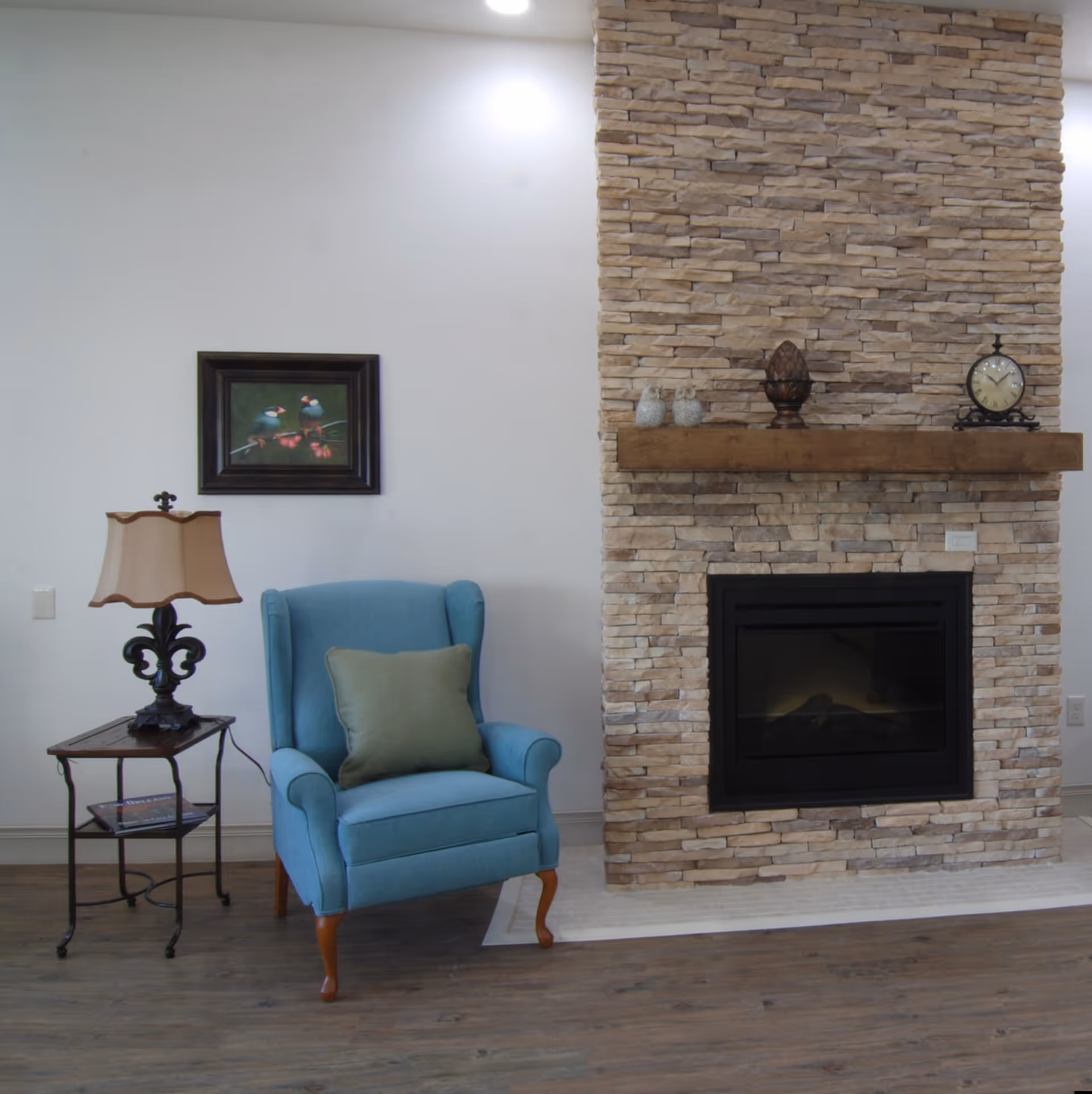 A cozy seating area with a blue armchair and side table beside a stone fireplace with a wooden mantel.