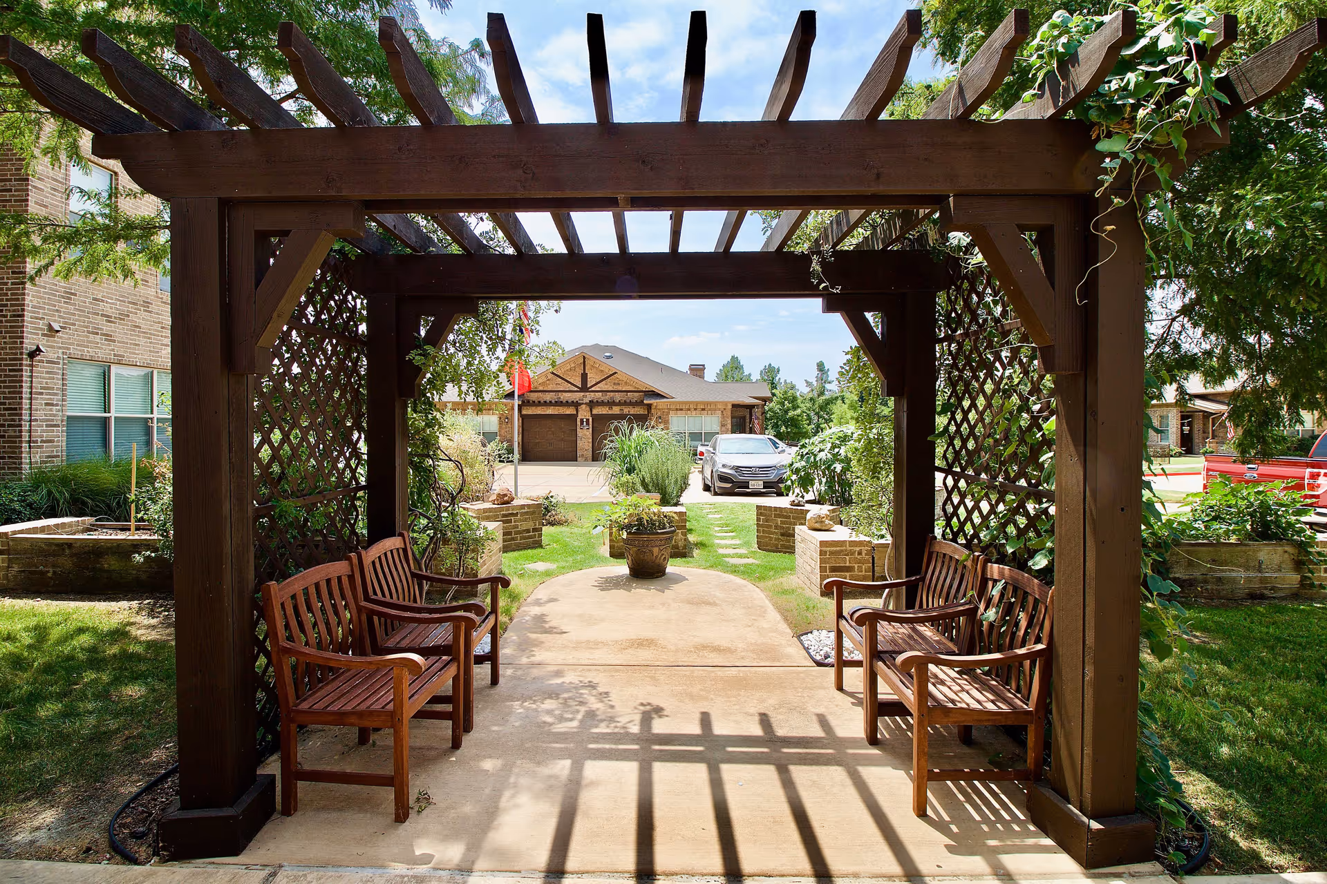 A wooden pergola with lattice sides and four wooden benches underneath, situated in a garden area with green grass and plants. In the background, there is a paved pathway leading to a building with a garage and parked cars visible.