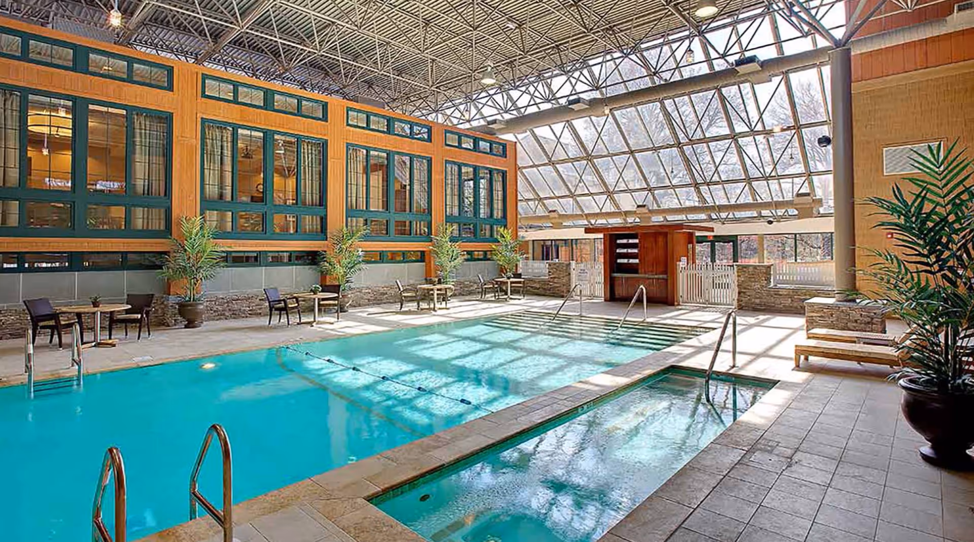 Indoor swimming pool area with a smaller hot tub adjacent to it, surrounded by tiled flooring and several tables with chairs. Large windows and a glass ceiling allow natural light to fill the space. Potted plants and wooden benches are placed around the pool area.