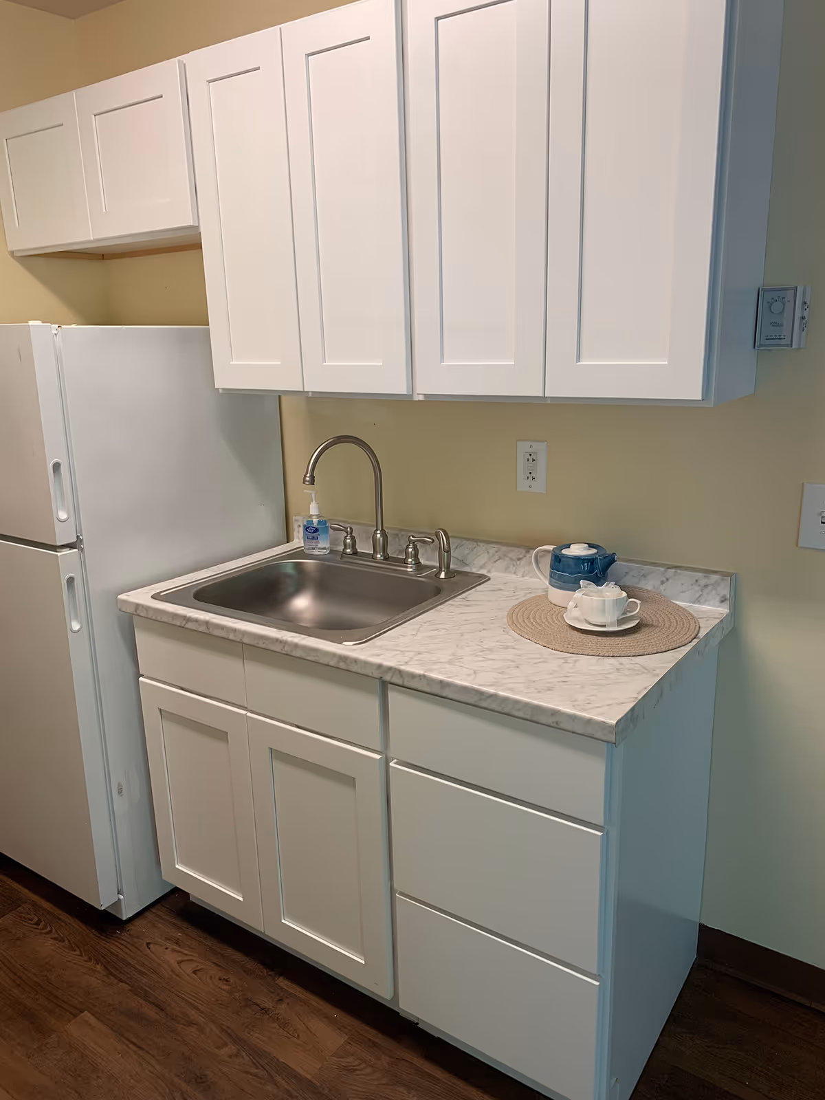 Compact kitchenette with white cabinets, a stainless steel sink and faucet, marble-look countertop, and a refrigerator.