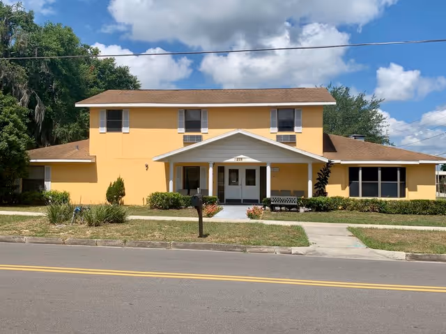 Front exterior view of a two-story yellow building with a brown roof, white trim, and a covered entrance with double doors. There is a small garden with bushes and flowers in front, a bench to the right of the entrance, a mailbox near the sidewalk, and a clear blue sky with some clouds above.