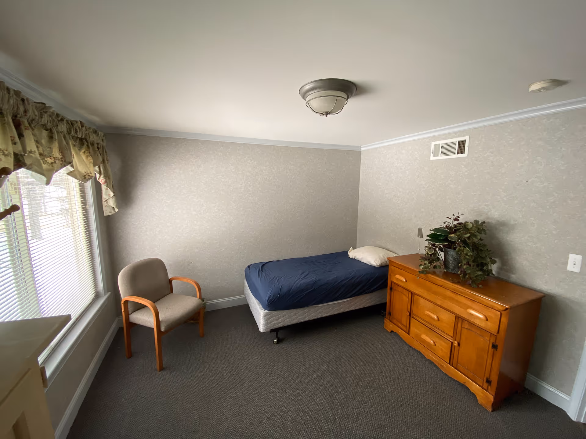 A small bedroom with a single bed covered with a dark blue blanket and a white pillow. Next to the bed is a wooden dresser with a potted plant on top. There is a beige armchair near a window with floral curtains and blinds. The walls are light gray with a subtle pattern, and the ceiling has a simple light fixture.