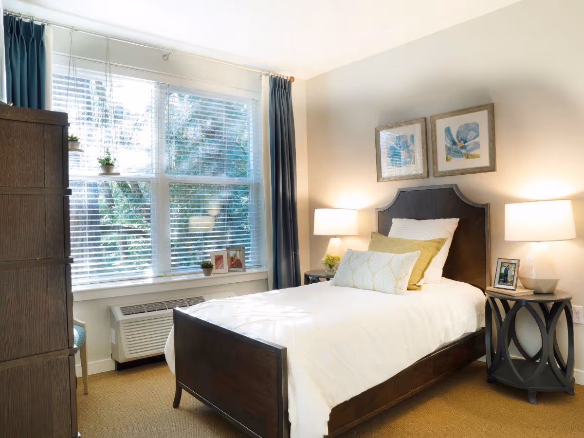 A bright and cozy bedroom with a single dark wood bed featuring white and green pillows and bedding. Two bedside tables with lamps and framed photos flank the bed. Large windows with white blinds and blue curtains allow natural light to fill the room. A dark wood dresser is visible on the left side.