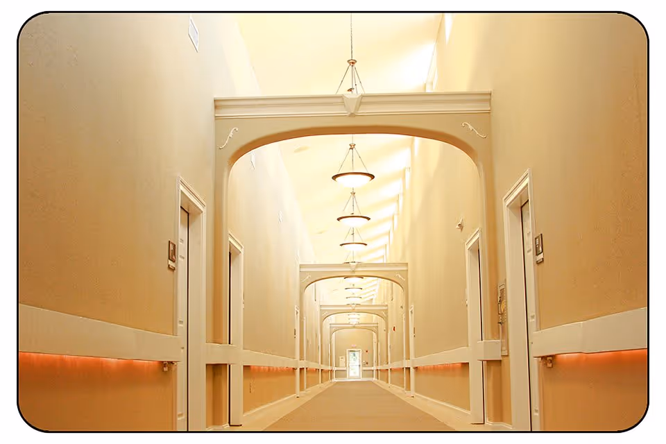 A long, well-lit hallway in a senior living community with beige walls, white doors on both sides, decorative arches, and ceiling lights evenly spaced along the corridor.