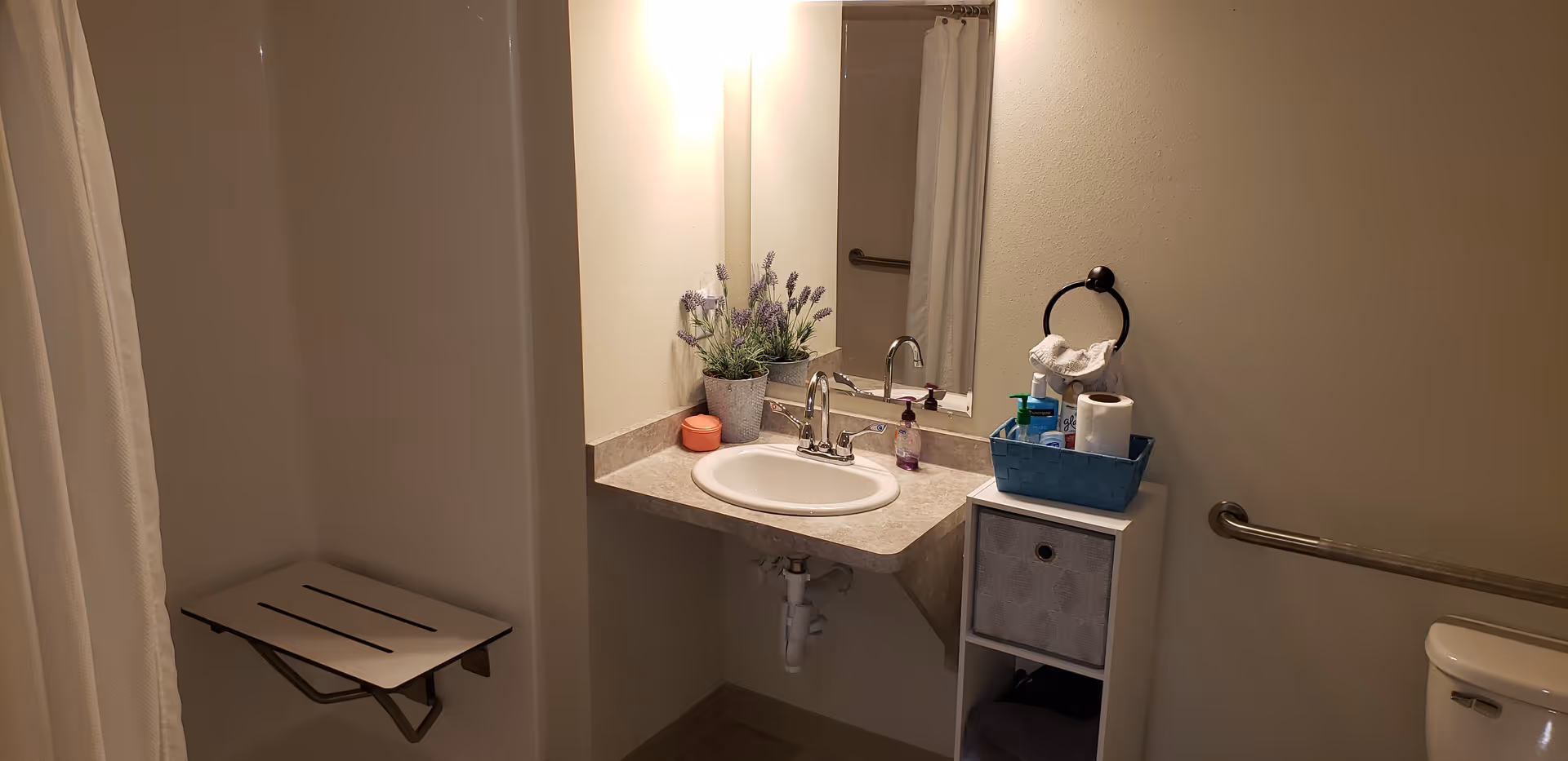 A bathroom with a wall-mounted foldable shower seat, a sink with a countertop, a mirror above the sink, a small potted plant, soap dispenser, and a storage unit holding toiletries and a roll of paper towels. There is a towel ring with a white towel and a grab bar next to the toilet.