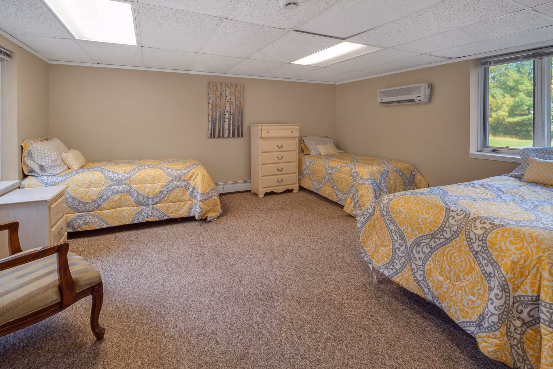 A bedroom with three single beds, each covered with yellow and gray patterned bedspreads. The room has beige walls, a carpeted floor, a window showing greenery outside, a small chest of drawers between two beds, a wooden chair with striped upholstery, and a wall-mounted air conditioning unit.