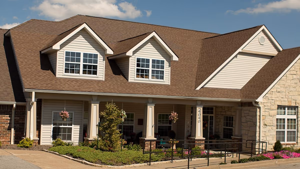 Front exterior view of a senior living facility building with beige siding, stone accents, and a brown shingled roof. The building has a covered porch with columns, hanging flower baskets, and a wheelchair ramp. The address number 4040 is visible on one of the columns. There are landscaped bushes and flowers in front of the building under a partly cloudy sky.